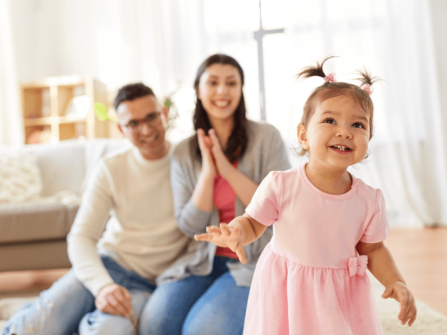 Two parents cheering on a baby walking in the foreground